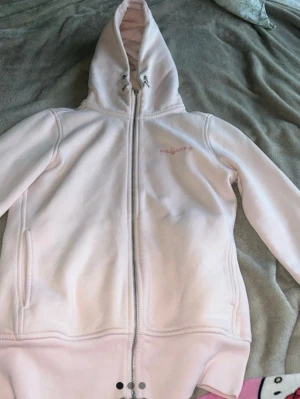 Ljusrosa fleecehoodie från Endurance - Mysig ljusrosa hoodie i fleece från Sail rancing med dragkedja framtill och broderad logga på bröstet. Hoodien har justerbar huva med snören och två sidofickor. Perfekt för chill dagar eller när du vill ha det extra bekvämt.