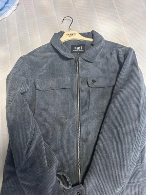 Grå manchester overshirt från Ami - Säljer en snygg grå overshirt från Ami i mjuk manchester. Jackan har två bröstfickor med lock, dragkedja framtill och klassisk krage. Perf cross-over mellan skjorta och jacka, med diskret Ami-logga på bröstet. Perfekt routing-plagg för lager på lager.