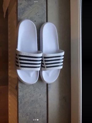 Vita Adidas badtofflor med svarta ränder - Säljer ett par klassiska vita  badtofflor med tre svarta ränder över foten. Tofflorna har en bred rem och är gjorda i syntetmaterial med räfflad sula för bra grepp. Perfekta för stranden eller gymmet.