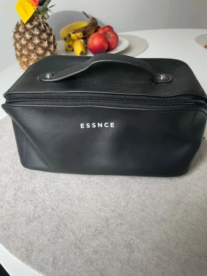 Svart necessär från ESSNCE - Stilren svart necessär från ESSNCE med dragkedja och praktiskt handtag. Väskan har ett rymligt fack och ett innerfack med dragkedja, perfekt för att organisera dina småsaker. Materialet är slätt och tåligt, med minimalistisk design och vit logotyp på framsidan. Har aldrig använd den ( endast varit liggande i garderoben).