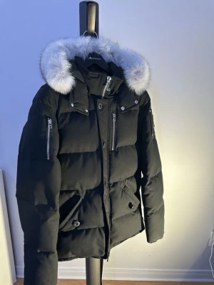 Svart dunjacka från Moose Knuckles - Snygg svart dunjacka från Moose Knuckles med pösig passform, silverfärgade dragkedjor och knappar samt stor huva med vit fuskpäls. Jackan har flera fickor och ikonisk logga på ärmen. Perfekt för kalla vinterdagar.