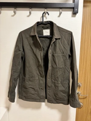 Olivgrön overshirt från A days march - Stilren olivgrön overshirt från Livid Jeans i kraftigt bomullstyg. Klassisk krage, tre stora utanpåfickor och knäppning framtill. Perfekt lager-på-lager-plagg med avslappnad passform och snygga detaljer.