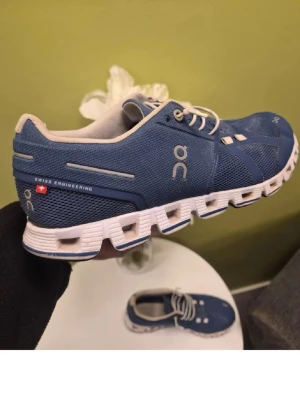 Blå On Cloud sneakers - Snygga blå On Cloud sneakers med vit sula och grå detaljer. Skorna har ventilerande meshmaterial och klassisk snörning. Perfekta för dig som gillar sportig stil och schweizisk design. Ikonisk On-logga på sidan och hälen.