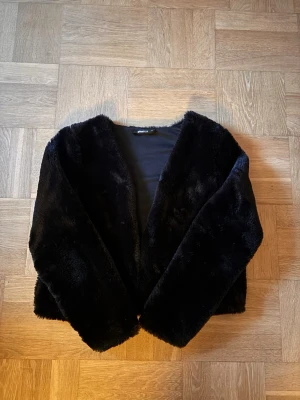 Svart kort pälsjacka - Snygg svart pälsjacka i fuskpäls med kortare modell och öppen front. Jackan har en mjuk och fluffig känsla, perfekt för att hålla värmen under kalla dagar