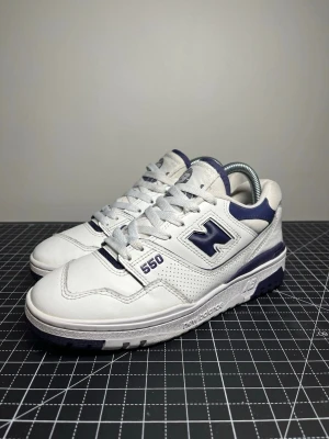 New Balance 550 - Klassiskas sneakers från New Balance, modell 550 i vit färg med lila detaljer. Retroinspirerad basketdesign med ren silhuett och perforerad läderovandel. Stabil sula och bekväm konstruktion som gör dem perfekta för vardaglig användning. Stilrena sneakers som passar bra till både casual och sportig stil.