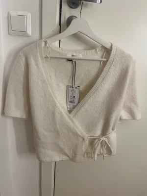 Vit omlottopp från Gina Tricot - Superfin vit omlottopp från Gina Tricot med kort ärm och knytning i sidan. Mjuk, stickad känsla och djup v-ringning. Perfekt att styla med jeans eller kjol för en trendig look. Aldrig använd med prislappen kvar - originalpris: 359 kr