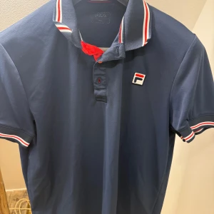 Marinblå piké från Fila - Snygg marinblå pikétröja från Fila med klassisk krage och knappar. Kragen och ärmsluten har vita och röda ränder som ger en sportig look. Fila-logga på bröstet. Perfekt för dig som gillar stilren och bekväm design. Passar bra till sommarens Tennis,Padel eller golf. Storlek M men passar S