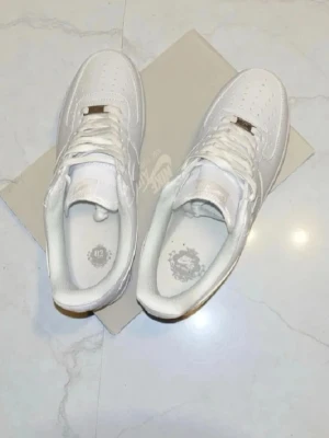 Vita Nike Air Force 1 sneakers - Klassiska helvita Nike Air Force 1 sneakers med lågt skaft och perforerad tå. Skorna har en stilren design med Nike-logga på sidan och plös, samt vita skosnören och sula. Perfekta för en clean och tidlös look.