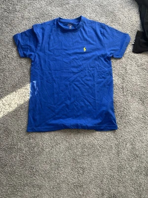 Blå t-shirt från Polo Ralph Lauren - Klassisk blå t-shirt från Polo Ralph Lauren i custom fit. T-shirten har korta ärmar och den ikoniska gula broderade loggan på bröstet. Tillverkad i mjuk bomull som känns skön mot huden. Perfekt till jeans eller shorts för en clean look. Pris kan diskuteras 