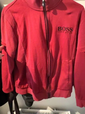 Röd zip hoodie från Hugo Boss - Snygg röd hoodie med dragkedja från Hugo Boss. Klassisk design med ribbade muddar, hög krage och logga broderad på bröstet. Tillverkad i mjukt bomullsmaterial som känns skönt mot huden. Perfekt för en avslappnad och stilren look.