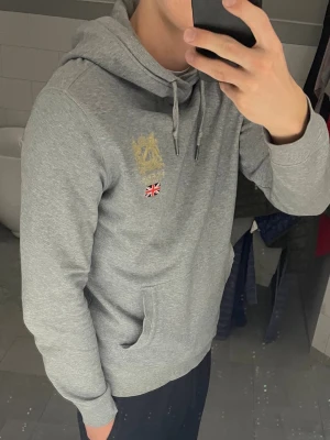 Grå hoodie med brodyr och ficka - Snygg grå hoodie från Morris med broderad logga i guld och brittisk flagga på bröstet. Klassisk känguruficka framtill och justerbar dragsko i huvan. Perfekt för en avslappnad och sportig stil.