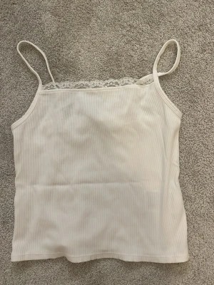 Vit ribbad linnetopp från H&M - Säljer en söt vit ribbad linnetopp från H&M med tunna axelband och spetsdetalj upptill. Perfekt att styla till jeans eller kjol. Materialet är mjuk bomull och toppen har en enkel, clean look med en feminin touch. storlek 146/152❤️❤️