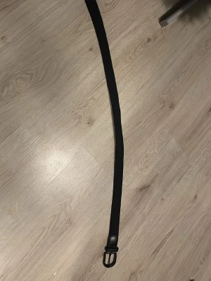 Svart flätat från JG - Snyggt svart bälte från JG med ett stilrent metallspänne i svart. Bältet har en klassisk design och passar perfekt till jeans eller byxor. Enkel att justera och ger en clean look till outfiten.