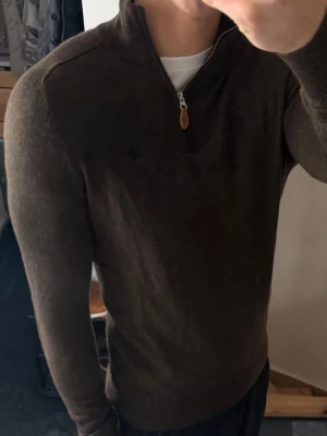 Brun  halvzip Morris - Mörkbrun stickad tröja med halvzip från Morris. Tröjan har ett diskret broderat emblem på bröstet och ribbade muddar vid ärmslut och nederkant. Perfekt för lager-på-lager och enkel att matcha med jeans eller chinos. Lappen är lite av men påverkar inte användningen. Liten i storleken sitter som xs