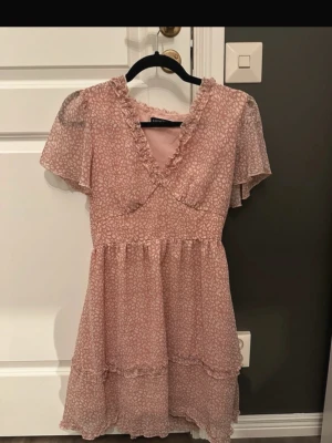 Ljusrosa blommig kortklänning - Supersöt ljusrosa kortklänning från Gina Tricot med småblommigt mönster, volangdetaljer och v-ringning. Klänningen har korta, transparenta ärmar och omlott-look upptill. Perfekt för sommarens alla tillfällen!