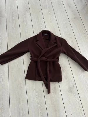 Mörkbrun kappa med knytskärp - Stilren mörkbrun kappa från missäy med klassisk krage och knytskärp i midjan. Kappan har raka ärmar och en enkel, tidlös design som passar perfekt till höst och vår. Materialet känns mjukt och ger en snygg siluett. Köpt för ca 500. Pris går att diskutera. Aldrig använd pga fel storlek.