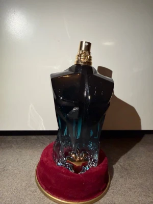 Jean Paul Gaultier Le Male parfym - Ikonisk parfymflaska från Jean Paul Gaultier formad som en manlig torso i blått och svart glas med guldfärgad spraytopp. Flaskan har en unik design och står på en rund röd sammetsbas med guldkant. Perfekt för dig som gillar snygga detaljer på parfymhyllan. KVITTO FINNS 
