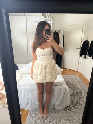 Krämvit axelbandslös kortklänning - Supersöt axelbandslös kortklänning i krämvit färg med draperad överdel och volangliknande struktur på kjolen. Perfekt för dig som vill sticka ut med en romantisk look. Kjolen har ett unikt rosmönster och klänningen har en figurnära passform upptill.