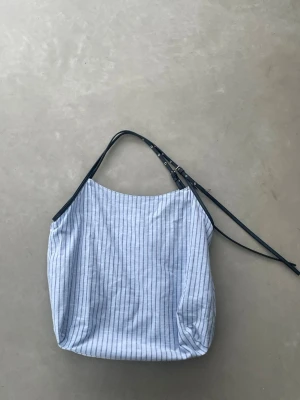 Blå randig tygväska från Zara - Snygg tygpåse från Zara i blått randigt tyg med svarta justerbara axelband i konstläder. Väskan har en mjuk, rymlig form och ett innerfack med dragkedja. Perfekt för dig som gillar en avslappnad men trendig stil.