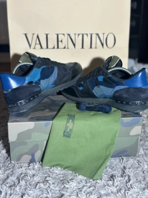 Blå Valentino rockrunners - Snygga sneakers från Valentino med blå och svarta camouflage-detaljer. Skorna har svarta snören, svarta nitar på sulan och blå häl. Materialmix av skinn och mocka ger en lyxig känsla. Perfekta för dig som vill sticka ut med stil. Og ingår ej