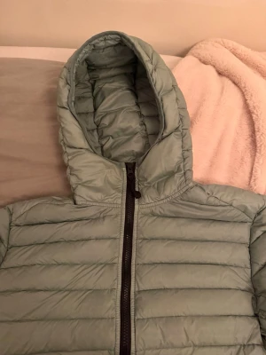 Grön dunjacka från Stone Island - Köpt 8000 kr i Frankrike Snygg ljusgrön dunjacka från Stone Island med klassisk patch på ärmen. Jackan har huva, dragkedja framtill och quiltad design. Perfekt för kalla vinterdagar och riktigt trendig stil.