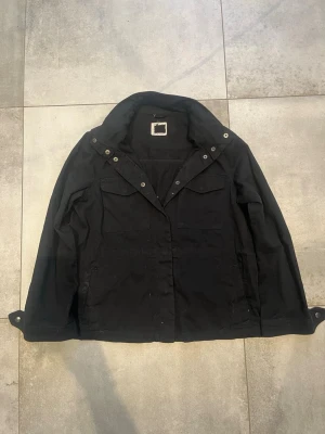 Jacka  - Stilren svart vindjacka med klassisk krage och tryckknappar framtill. Jackan har två bröstfickor med lock och två sidofickor. Långärmad modell med justerbara ärmslut och diskreta detaljer. Perfekt för dig som gillar enkel och clean stil.