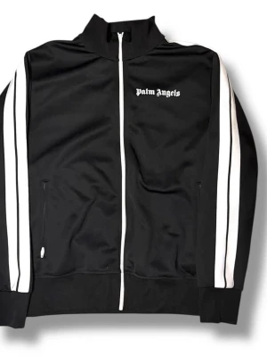 Svart Palm Angels zip hoodie - Snygg svart hoodie från Palm Angels med vit dragkedja och vita dubbla ränder längs ärmarna. Märkets logga i vitt på bröstet. Tillverkad i mjukt och stretchigt material, perfekt för en streetwear-look. Två fickor framtill och ribbade muddar vid ärmslut och nederkant.