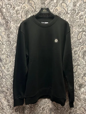 Svart sweatshirt från Stone Island - Svart sweatshirt från Stone Island med rund halsringning och diskret logopatch på bröstet. Tröjan har en clean look, ribbade muddar och är tillverkad i mjukt material som känns skönt mot huden. Finns i flera storlekar, skriv önskad storlek i samband med order. 