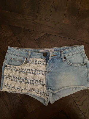 Ljusblå jeansshorts med spetsdetalj - Säljer ett par ljusblå jeansshorts från OshKosh med snygg spetsdetalj i vitt på ena framsidan. Shortsen har råa kanter, klassiska fickor och bälteshällor. Perfekta för sommaren och ger en cool, avslappnad vibe. Står att de är i storlek 152 vilket jag tycker är konstigt då midjemåttet är 39cm så skulle säga mer xs!