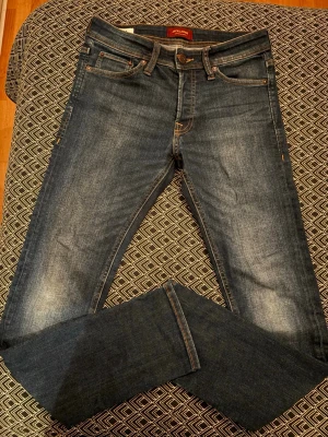 Blå slim fit jeans från Jack & Jones - Snygga blå slim fit jeans från Jack & Jones med klassisk femficksdesign och ljusa slitningar framtill. Jeansen har normalhög midja och är tillverkade i stretchigt denim för en bekväm passform. Perfekta för dig som gillar en smal siluett. Längd 32