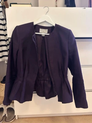 Mörklila kavaj med figursydd passform - Stilren mörklila kavaj med rundad krage och figursydd midja. Kavajen har långa ärmar, dold knäppning framtill och lätt utsvängd nederkant. Perfekt för en trendig och elegant look.