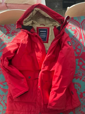 Röd parkas från Sebago med huva - Röd parkasjacka från Sebago med beige innerfoder och stor huva. Jackan har flera praktiska fickor, justerbar huva och Sebago-logga på ärmen. Perfekt för kalla dagar och har en klassisk amerikansk look. Materialet känns slitstarkt och varmt.