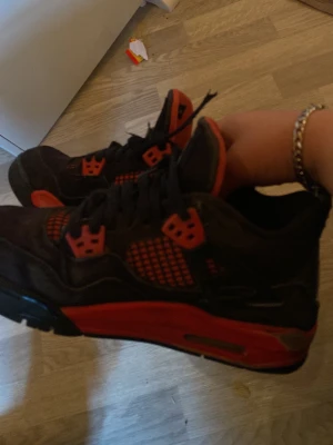 Svarta och röda Air Jordan 4 sneakers - Säljer ett par klassiska Air Jordan 4 sneakers i svart och rött med meshdetaljer och ikoniska Jumpman-loggan på hälen. Skorna har svarta skosnören, röda detaljer på sidorna och en synlig luftbubbla i sulan. Perfekta för dig som gillar streetwear och snygg design.