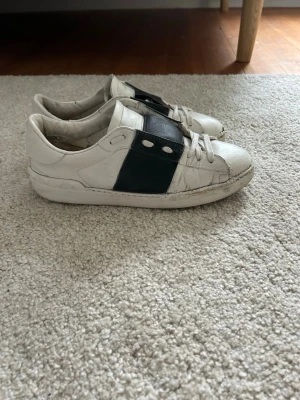 Valentino vita sneakers med svart detalj - Snygga vita sneakers från Valentino med bred svart läderdetalj över sidan och klassisk snörning. Skorna har en låg profil och är tillverkade i läder för en clean och stilren look. Kommer med originalkartong och dustbag.