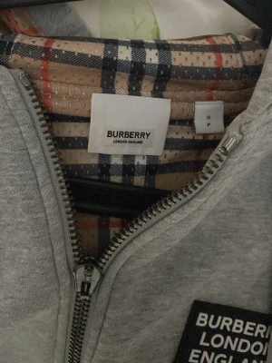 Grå zip hoodie från Burberry - Snygg grå hoodie från Burberry med dragkedja, huva och fickor framtill. Klassisk Burberry-rutig insida och patch med logga på bröstet. Perfekt för dig som gillar stilrena och exklusiva plagg med streetwear-vibe.