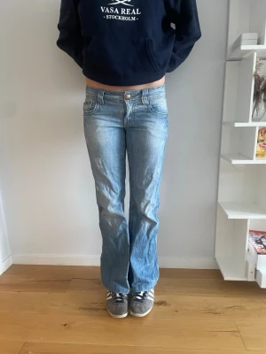 Blå lågmidjade bootcut jeans med slitningar - Säljer ett par ljusblå bootcut jeans med slitningar och tvättade detaljer. Jeansen  dekorativa sömmar på bakfickorna. Perfekta för en avslappnad och trendig look.