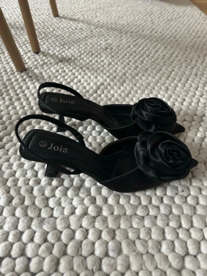 Svarta pumps med rosdetalj  från Joia - Stilrena svarta pumps från Joia med spetsig tå och låg klack. Skorna har en elegant rosdetalj i satin framtill och öppen häl med smal rem. Endast använda en gång