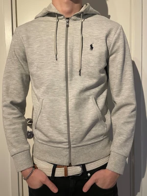 Polo Ralph Lauren zip hoodie   - Snygg ljusgrå hoodie med dragkedja från Polo Ralph Lauren. Klassisk design med huva och dragsnören, två fickor framtill och den ikoniska loggan broderad på bröstet. Tillverkad i mjukt bomullsmaterial som är skönt att ha på sig.