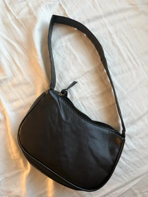 Svart axelremsväska i skinnimitation - Snygg svart handväska i skinnimitation med axelrem och dragkedja upptill. Väskan har en mjukt rundad form och minimalistisk design som passar till många olika stilar. Perfekt storlek för det viktigaste och enkel att matcha.