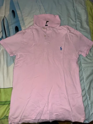 Ljusrosa pikétröja från Ralph Lauren - Säljer en ljusrosa pikétröja från Ralph Lauren med klassisk krage och två knappar framtill. Tröjan har korta ärmar och den ikoniska blå logotypen broderad på bröstet. Tillverkad i mjukt bomullsmaterial som andas.