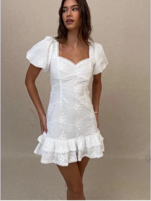 Vit kort klänning med puffärm - Söt vit kort klänning med puffärmar, broderade blommor och volangkant. Klänningen har knytning i ryggen och dekorativa pärlor längs urringningen. Perfekt för sommaren och studenten!