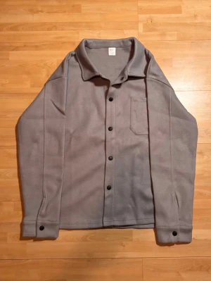 Grå overshirt - Säljer en stilren grå overshirt i mjuk fleece med klassisk krage, svarta knappar och en bröstficka. Perfekt lager på lager-plagg med avslappnad passform och långa ärmar med knapp vid ärmslut. Enkel och clean look som funkar till det mesta. 