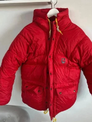 Röd dunjacka från Fjällräven - Riktigt fet röd dunjacka från Fjällräven med huva och gula detaljer på snörena. Jackan har två stora fickor framtill med knappar och klassisk Fjällräven-logga på bröstet. Perfekt för kalla vinterdagar och riktigt varm. finns en liten skromma på ena armen men inget man märker o går nå lätt att laga , men där av det nedsatta priset 