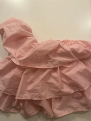 Rosa volangig oneshoulder topp Anielle - Supersöt rosa topp från Anielle med volanger och oneshoulder-design. Toppen är axelbandslös på ena sidan och har en luftig, flowy känsla. Perfekt för dig som vill sticka ut med en feminin och trendig look.