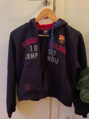 Mörkblå FC Barcelona hoodie - Snygg mörkblå hoodie från FC Barcelona med dragkedja, huva och fickor framtill. Tryck med klubbens logga och texten 'FCBARCELONA 1899 CAMP NOU' samt röd insida i huvan. Perfekt för dig som älskar fotboll och vill visa ditt lagstöd.