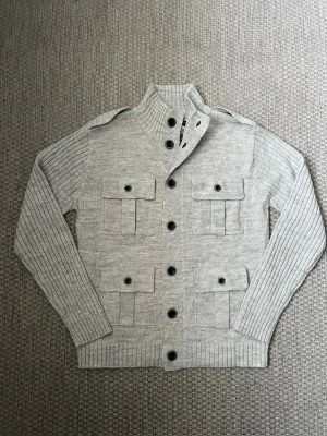 Ljusgrå stickad cardigan med fickor - Snygg ljusgrå stickad cardigan med hög krage och svarta knappar. Fyra stora fickor framtill med lock och knappstängning. Ribbstickade ärmar och muddar ger en cool och modern look. Perfekt för lager-på-lager-stil.