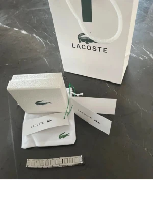 Silverfärgad klocklänk från Lacoste - Snygg silverfärgad klocklänk från Lacoste med klassisk länkdesign. Kommer med originalförpackning, tygpåse och tags. Perfekt för dig som vill uppgradera din klocka med en stilren och tidlös accessoar från ett välkänt märke.