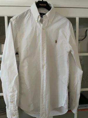 Vit skjorta från Ralph Lauren Slim Fit - Klassisk vit skjorta från Ralph Lauren i slim fit-modell. Skjortan har lång ärm, knappar framtill och den ikoniska broderade loggan på bröstet. Perfekt till både jeans och chinos för en clean och stilren look.