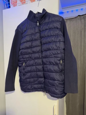 Mörkblå cardiganjacka från Moncler - Snygg mörkblå cardiganjacka från Moncler med quiltad framsida och stickade ärmar. Jackan har hög krage, två dragkedjefickor och Moncler-logga på ärmen. Perfekt för lager på lager och en stilren look. Bara att skriva vid frågor.