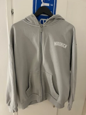 Grå zip hoodie från Hoodrich - Snygg grå hoodie med dragkedja från Hoodrich. Hoodien har huva, två stora fickor framtill och ribbade muddar vid ärmslut och nederkant. Stort tryck med HOODRICH på bröstet. Perfekt för en avslappnad och street-inspirerad stil.